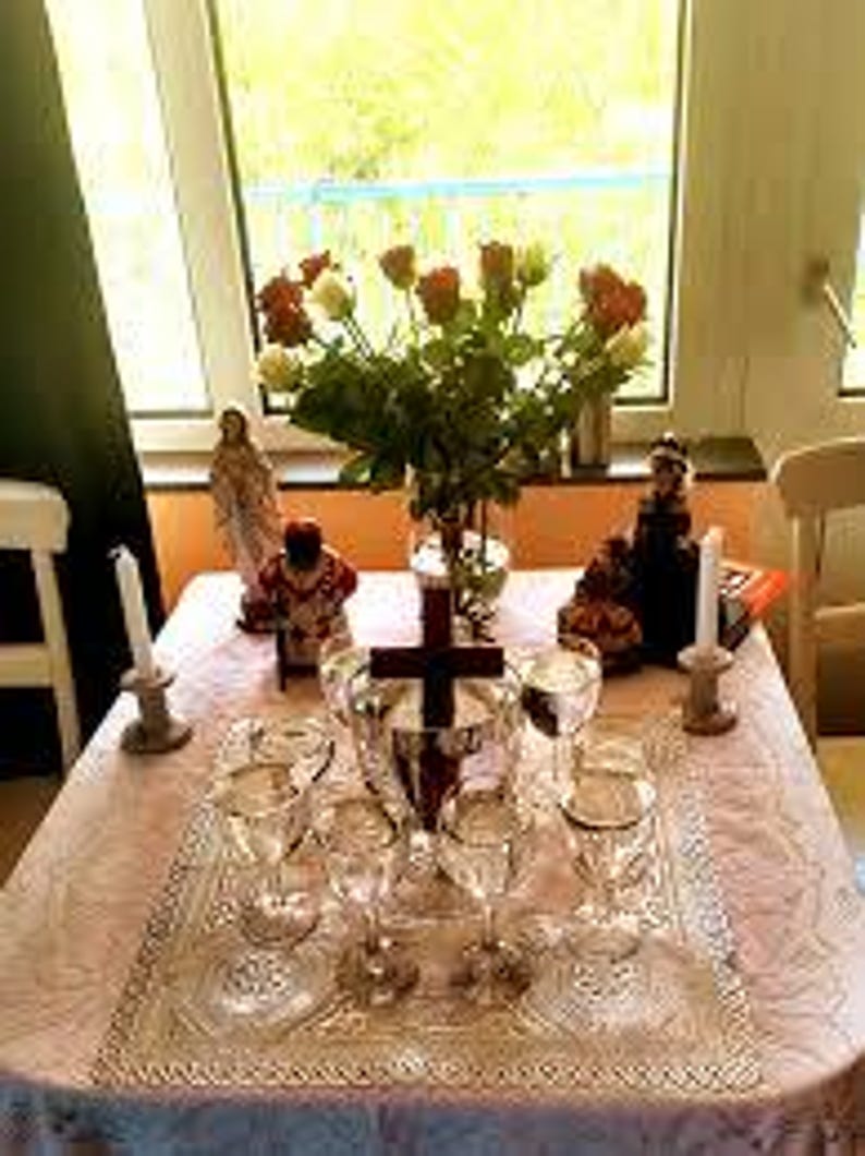 May include: A white tablecloth with a lace pattern covers a table with a wooden cross, six clear glass goblets, two white candles, and three religious figurines. The figurines are a woman in a white dress, a man in a red and gold robe, and a woman in a red and black dress. A bouquet of red and white roses is in a clear glass vase.