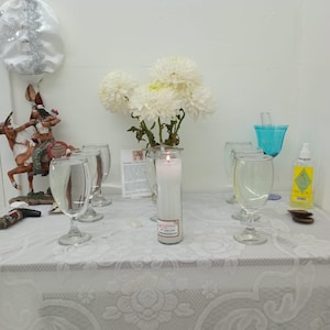 May include: A white table cloth with a lace pattern covers a table with a white candle, four clear glass cups, a small figurine, and a bouquet of white flowers.