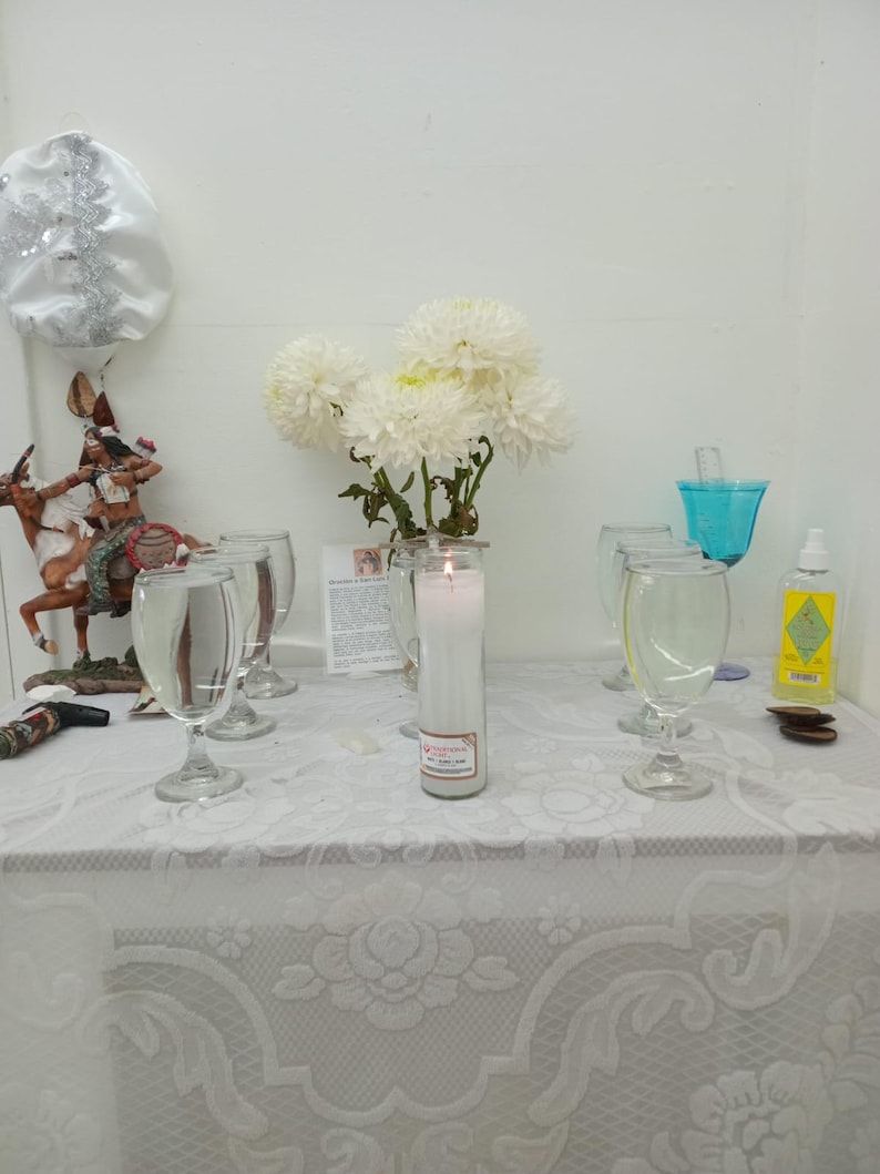 May include: A white table cloth with a lace pattern covers a table with a white candle, four clear glass cups, a small figurine, and a bouquet of white flowers.