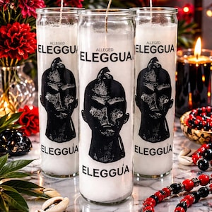 May include: Three white candles in glass jars with the text "ALLEGED ELEGGUA" and a black graphic. The candles are surrounded by red flowers, beads, and seashells. A lit candle is in the background.