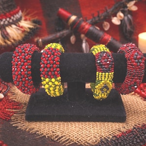 May include: Four beaded bracelets in red, yellow, and black are displayed on a black stand. The bracelets are made of small beads and have intricate patterns. The background includes candles, decorative items, and beaded necklaces.