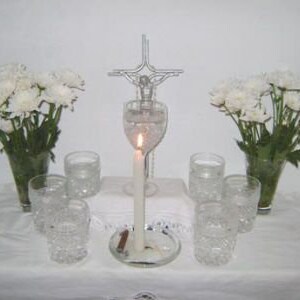 May include: A white table with a silver cross, a lit candle in a glass vase, and white flowers in two vases. There are six clear glass cups on the table.