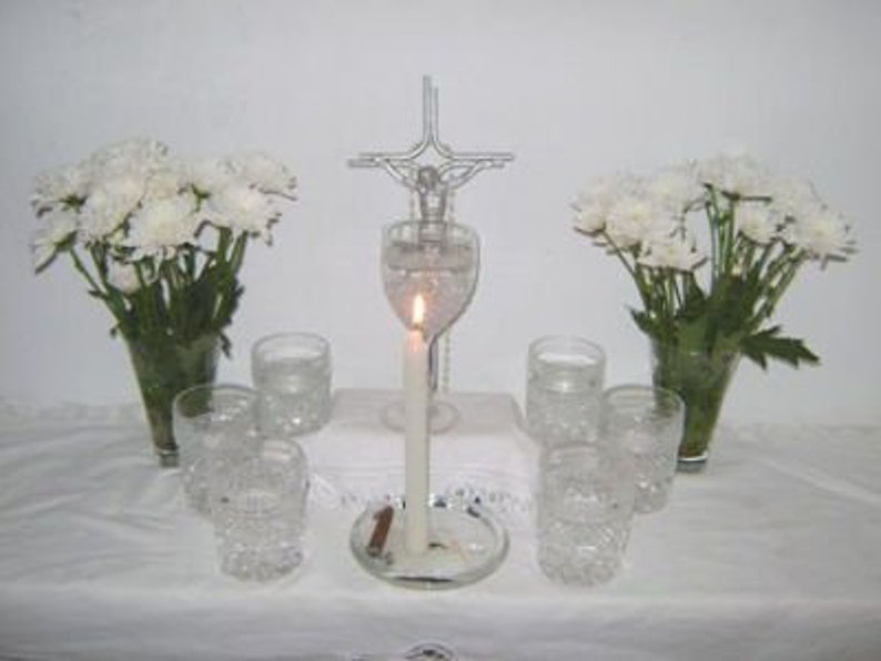 May include: A white table with a silver cross, a lit candle in a glass vase, and white flowers in two vases. There are six clear glass cups on the table.