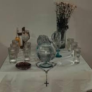 May include: A white tablecloth with a variety of glass items, including a pitcher, glasses, and a bowl filled with water. A rosary with a silver cross is in the center of the table. A bouquet of dried white flowers is in a glass vase in the background.