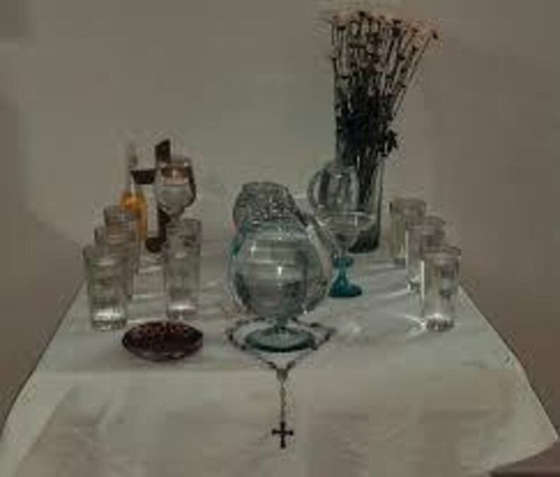 May include: A white tablecloth with a variety of glass items, including a pitcher, glasses, and a bowl filled with water. A rosary with a silver cross is in the center of the table. A bouquet of dried white flowers is in a glass vase in the background.