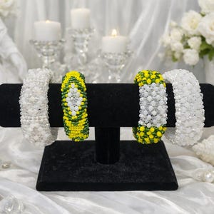 May include: Four beaded bracelets displayed on a black stand. Two are white, and two are yellow and green. The bracelets are made of small beads and are arranged in a row. The background includes candles, flowers, and decorative items.