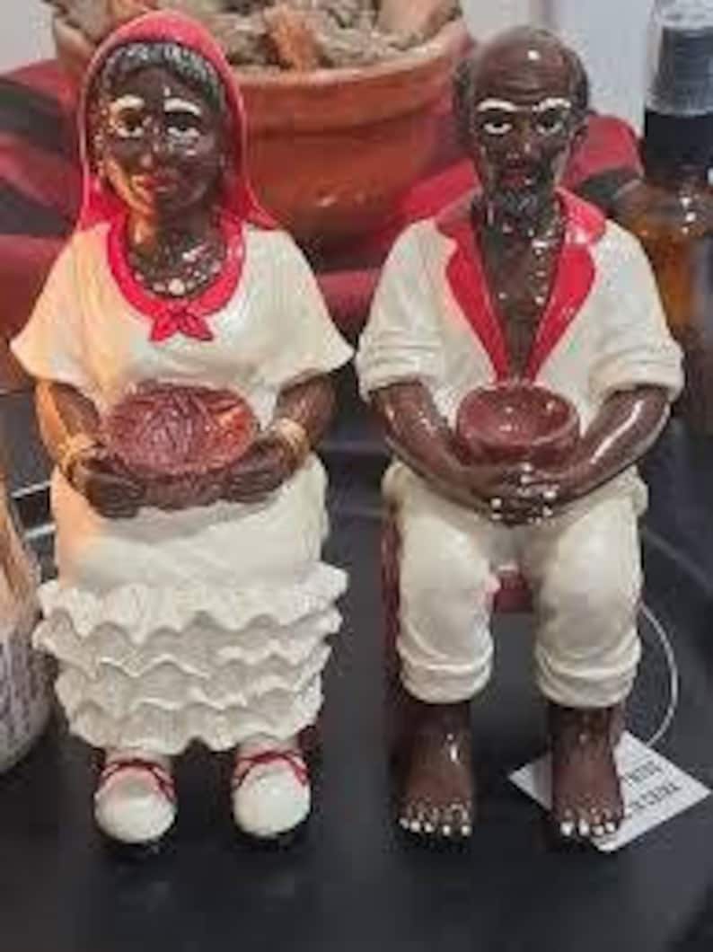 May include: A pair of small, hand-painted figurines depicting a man and woman. The woman is wearing a white dress with a red shawl and holding a basket. The man is wearing white pants and a white shirt with a red sash and holding a bowl. Both figurines have brown skin and black hair.