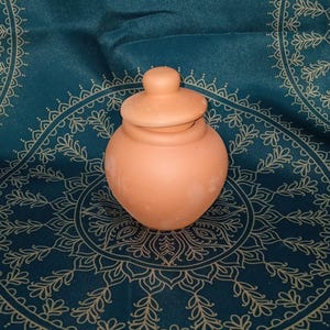 May include: A small, terracotta-colored jar with a lid. The jar has a rounded body and a small, knob-like handle on the lid. The jar is sitting on a teal-colored fabric with a gold-colored floral pattern.