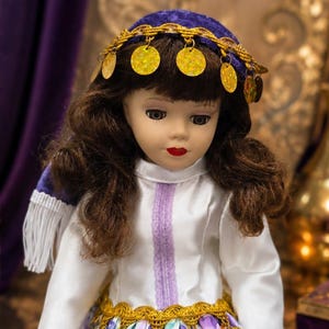 May include: A doll with brown hair wearing a purple velvet headpiece adorned with gold dangling coins. The doll features a white long-sleeved top with a vertical lavender stripe, a gold braided waistband, and a white fringe detail on the side.