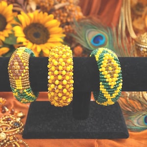 May include: Three beaded bracelets in yellow, green, and brown are displayed on a black stand. The bracelets are adorned with small beads, creating a textured surface. Sunflowers and peacock feathers are in the background.