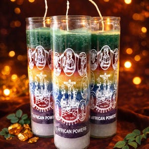 May include: Three glass candles with layered colors of green, yellow, orange, blue, and purple. Each candle has a white label with illustrations and the text "AFRICAN POWERS". The candles are on a dark red surface with gold accents and a blurred background.