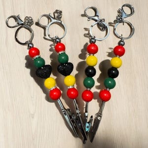 May include: Four beaded keychains with silver-tone clips. Each keychain features a series of colorful beads in red, green, yellow, and black, with a black heart-shaped bead on two of them. The keychains are on a light wood surface.