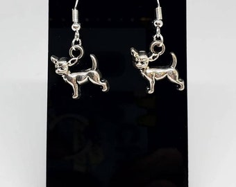 May include: Silver-colored Chihuahua dog earrings. The earrings are displayed on a black stand against a white background. The Chihuahua charms are detailed and have a hook closure.