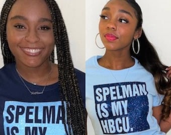 May include: Two women wearing blue and light blue t-shirts with the text "Spelmen is my HBCU." The woman on the left is wearing a navy blue t-shirt with light blue glitter text. The woman on the right is wearing a light blue t-shirt with dark blue glitter text. Both women are wearing jeans.