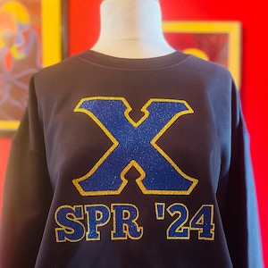 May include: Navy blue sweatshirt with a large blue and gold letter "X" and the text "SPR '24" in gold. The sweatshirt is displayed on a mannequin against a red background. The design is centered on the chest.