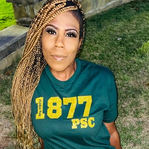 May include: A dark green t-shirt with gold glitter text that reads "1877 PSC" and "My HBCU". The person is wearing blue jeans with ripped knees and a brown belt with a gold buckle.