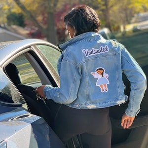 May include: A light blue denim jacket with a white patch that says "Undaunted" and a cartoon illustration of a girl in a white dress. The jacket has distressed details.