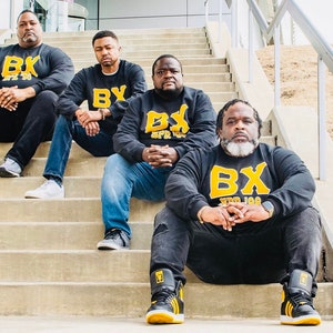 May include: Four men wearing black sweatshirts with yellow lettering that says "BX SPR 100" are sitting on a set of concrete stairs. The men are all smiling and looking at the camera.
