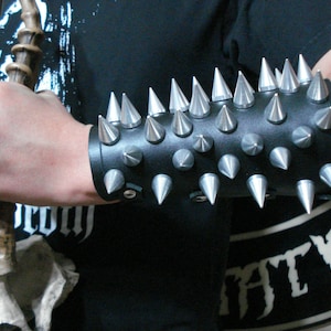 May include: A black leather wrist cuff with silver metal spikes. The cuff is worn on a person's left arm.