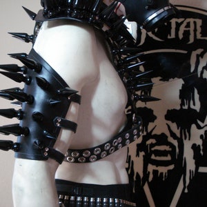 May include: Black leather arm armor with spikes and straps. The armor is worn on a mannequin and has a metal buckle. The background is a black and white image of a skull with the word "METAL" above it.