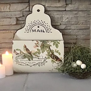May include: Cream-colored metal mail holder with a decorative bird and floral design. The word "MAIL" is printed at the top. Two lit candles and a small bird's nest with eggs are also present.