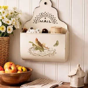 May include: Cream-colored metal mail holder with a decorative bird and butterfly design. The top features the word "MAIL" in black. The holder is mounted on a white paneled wall, with a wooden bowl of apples and a basket of flowers nearby.
