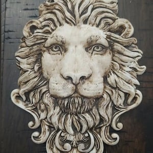 May include: A detailed, off-white lion's head sculpture with a textured mane and facial features. The sculpture has a regal appearance, with intricate details in the fur and decorative elements. The background is a dark, wooden surface.