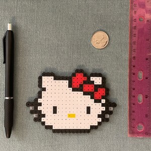 Pu&ograve; includere: Un personaggio Hello Kitty bianco fatto di perline perler con un fiocco rosso. Il personaggio &egrave; circondato da un bordo nero.