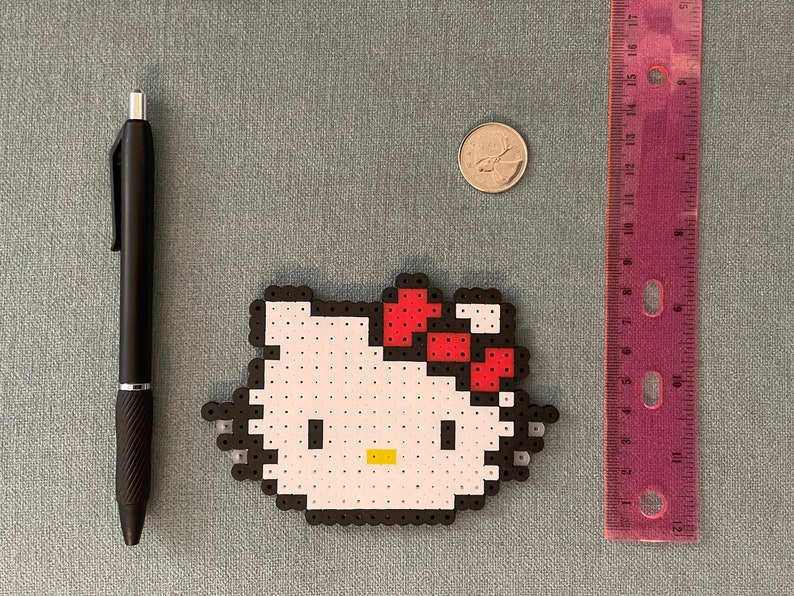 Pu&ograve; includere: Un personaggio Hello Kitty bianco fatto di perline perler con un fiocco rosso. Il personaggio &egrave; circondato da un bordo nero.