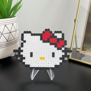 Pu&ograve; includere: Una figurina di Hello Kitty in pixel art bianca e nera con un fiocco rosso sulla testa. La figurina &egrave; su un supporto in plastica trasparente.