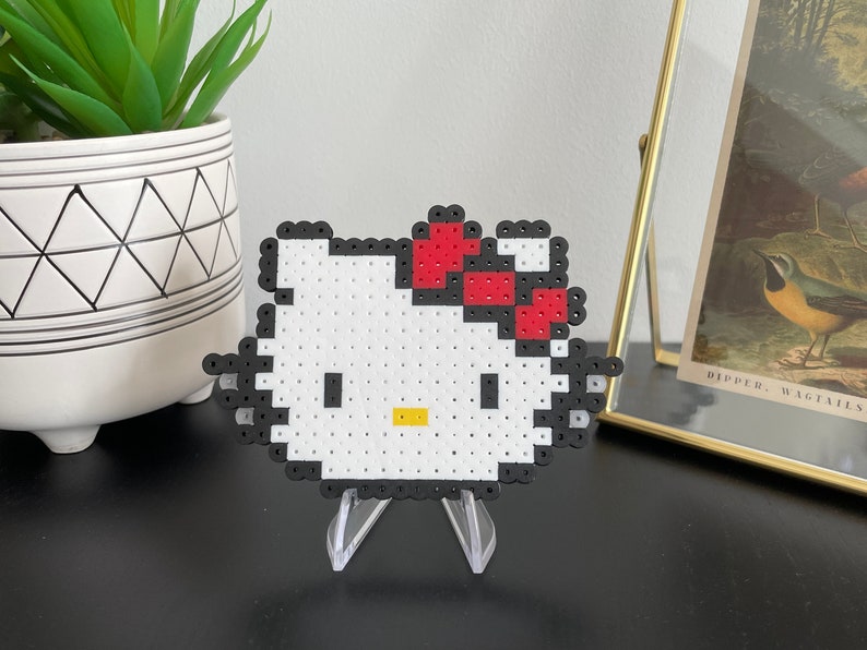 Pu&ograve; includere: Una figurina di Hello Kitty in pixel art bianca e nera con un fiocco rosso sulla testa. La figurina &egrave; su un supporto in plastica trasparente.