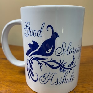 May include: White ceramic coffee mug with a blue design of a bird on a branch. The mug has the text "Good Morning Asshole" in blue cursive lettering.