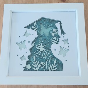 May include: A white frame with a cutout design of a person wearing a graduation cap and gown. The cutout is filled with a green and white floral pattern and butterflies.