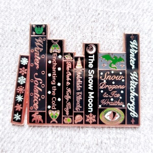 May include: A black and gold enamel pin depicting a stack of books with titles related to winter and magic. The titles include "Winter Solstice", "Enchanting the Cold", "The Oak & Holly Kings", "Yuletide Rituals", "The Snow Moon", "Snow Dragons & Ice Wraiths", and "Winter Witchcraft".