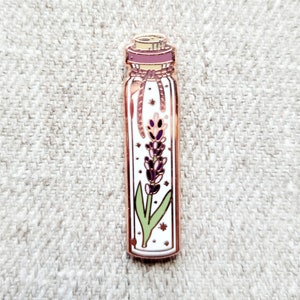 May include: A white enamel pin with a gold outline depicting a glass bottle with a cork stopper and a purple flower inside. The bottle has a string tied around the neck and stars are visible inside the bottle.