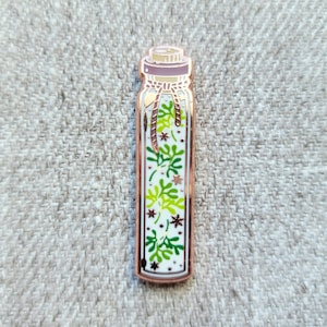 Könnte beinhalten: Eine goldfarbene Emaille-Anstecknadel mit einer Glasflasche, die mit grünen und weißen Blättern und kleinen weißen Sternen gefüllt ist. Die Flasche hat einen Korkverschluss und ein braunes Band, das um den Hals gebunden ist.