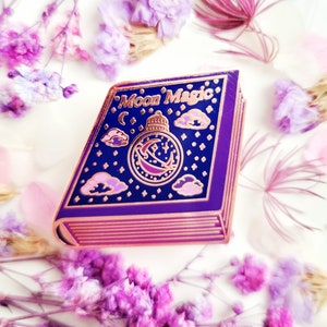Puede incluir: Un pin de esmalte de oro rosa con forma de libro cerrado con las palabras "Moon Magic" en la portada. El libro tiene una portada morada con un diseño de luna y estrellas doradas.