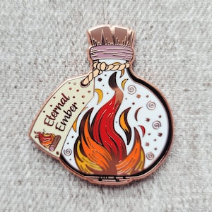 May include: A copper-coloured enamel pin shaped like a potion bottle with a white background and a red, orange, and yellow flame inside. The bottle has a tag that reads "Eternal Ember".