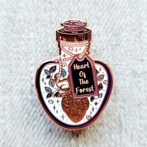 Puede incluir: Botella en forma de corazón de oro rosa con un tapón de corcho y una etiqueta que dice "Heart Of The Forest". La botella contiene un corazón estilizado con un árbol que crece de él.