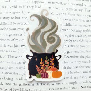 May include: A black cauldron with a green, orange, and brown floral design and a white and gray smoke plume rising from the top. The cauldron is filled with small orange, brown, and white pumpkins.