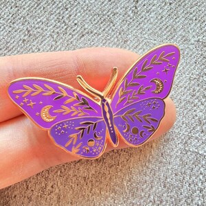 May include: Purple enamel butterfly pin with gold accents. The butterfly has intricate details of leaves, stars, and moons.