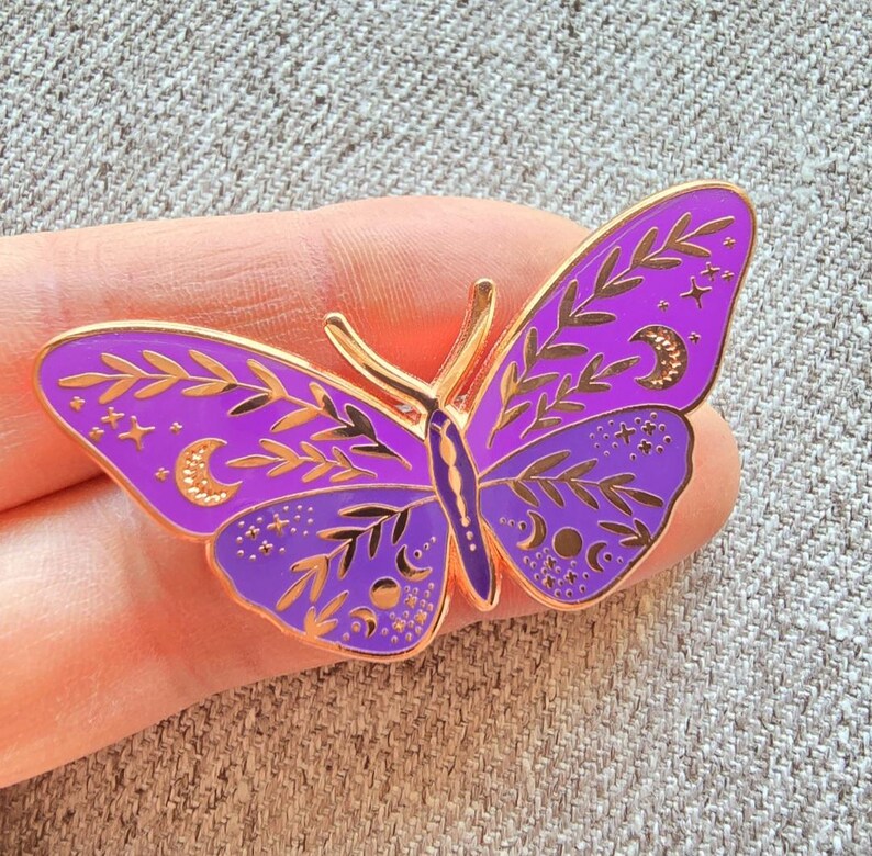 May include: Purple enamel butterfly pin with gold accents. The butterfly has intricate details of leaves, stars, and moons.
