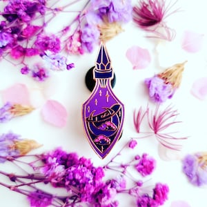 Puede incluir: Pin de esmalte morado y dorado de una botella de poción con una luna creciente y nubes en su interior. La botella tiene un tapón en forma de cristal.