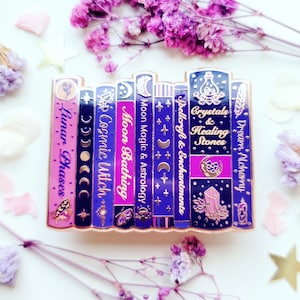 Puede incluir: Un conjunto de pines de esmalte que representan una pila de libros con títulos relacionados con la brujería y la magia. Los títulos incluyen "Fases lunares", "Bruja cósmica", "Baño de luna", "Magia lunar y astrología", "Hechicería y encantamientos", "Cristales y piedras curativas", y "Alquimia de los sueños".