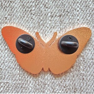 May include: Gold-colored metal butterfly pin with black metal clasps on the back.