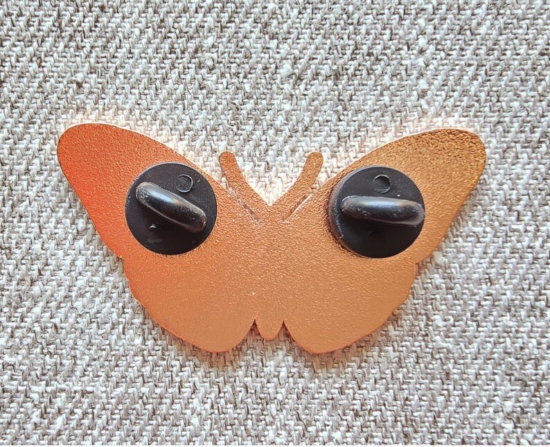 May include: Gold-colored metal butterfly pin with black metal clasps on the back.
