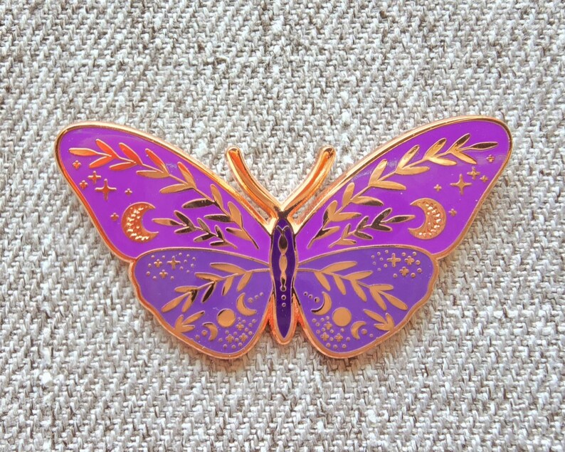 May include: A purple and gold butterfly enamel pin with intricate details, including leaves, stars, and moons.