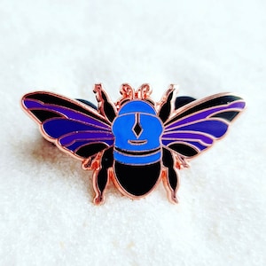 Puede incluir: Un pin de abeja de oro rosa con alas negras y moradas y un cuerpo azul. La abeja tiene una cara en forma de diamante.