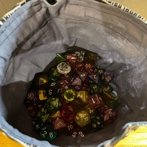 May include: A close-up of a grey fabric bag filled with a variety of colorful dice. The dice are made of clear plastic with different colored swirls and have numbers printed on them.