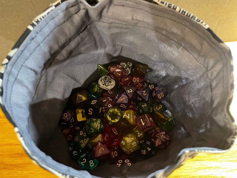 May include: A close-up of a grey fabric bag filled with a variety of colorful dice. The dice are made of clear plastic with different colored swirls and have numbers printed on them.